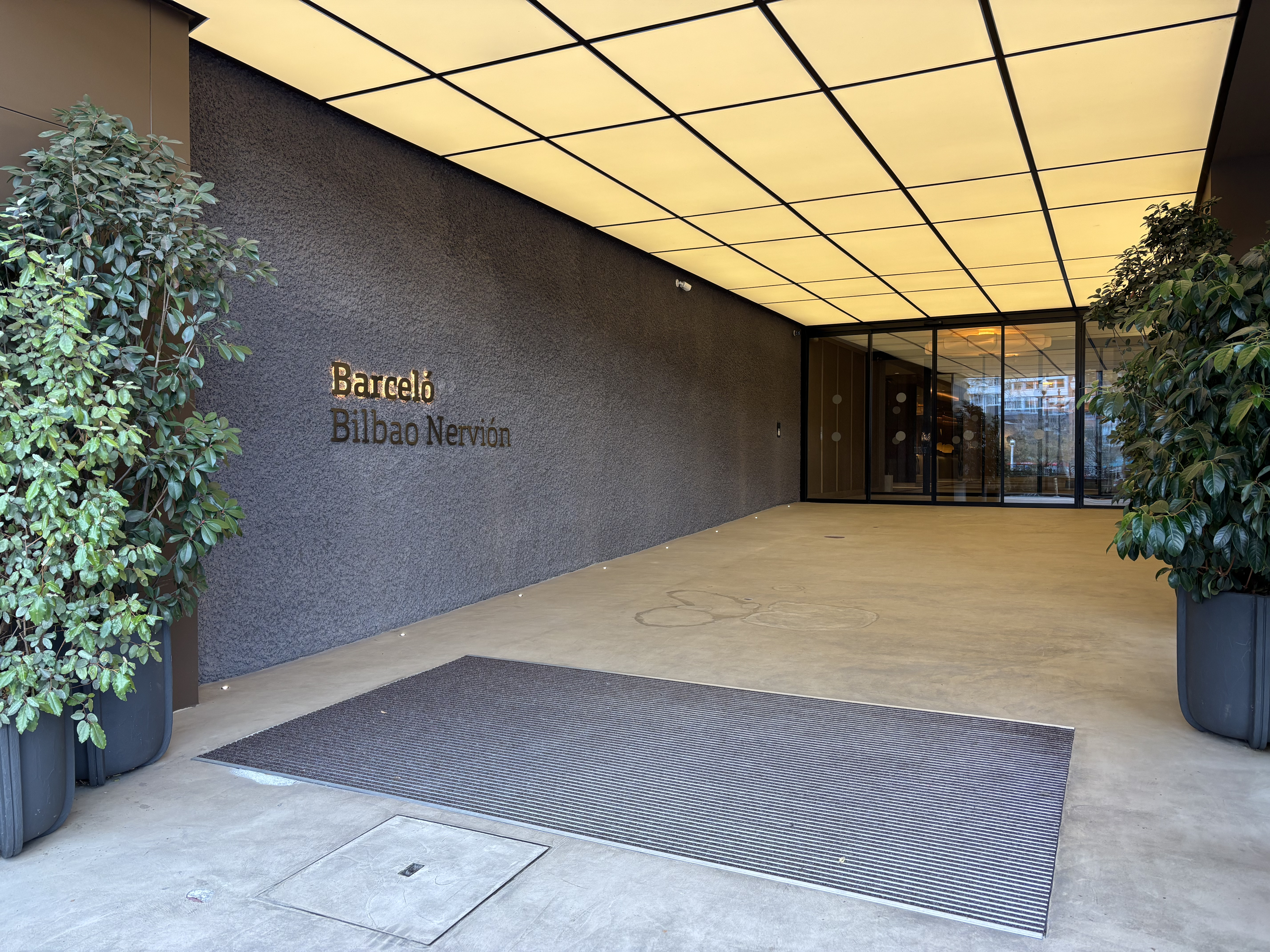 The image shows the entrance of a building. In the foreground, there is a gray entrance mat with slits. On either side of the door, there are two ...