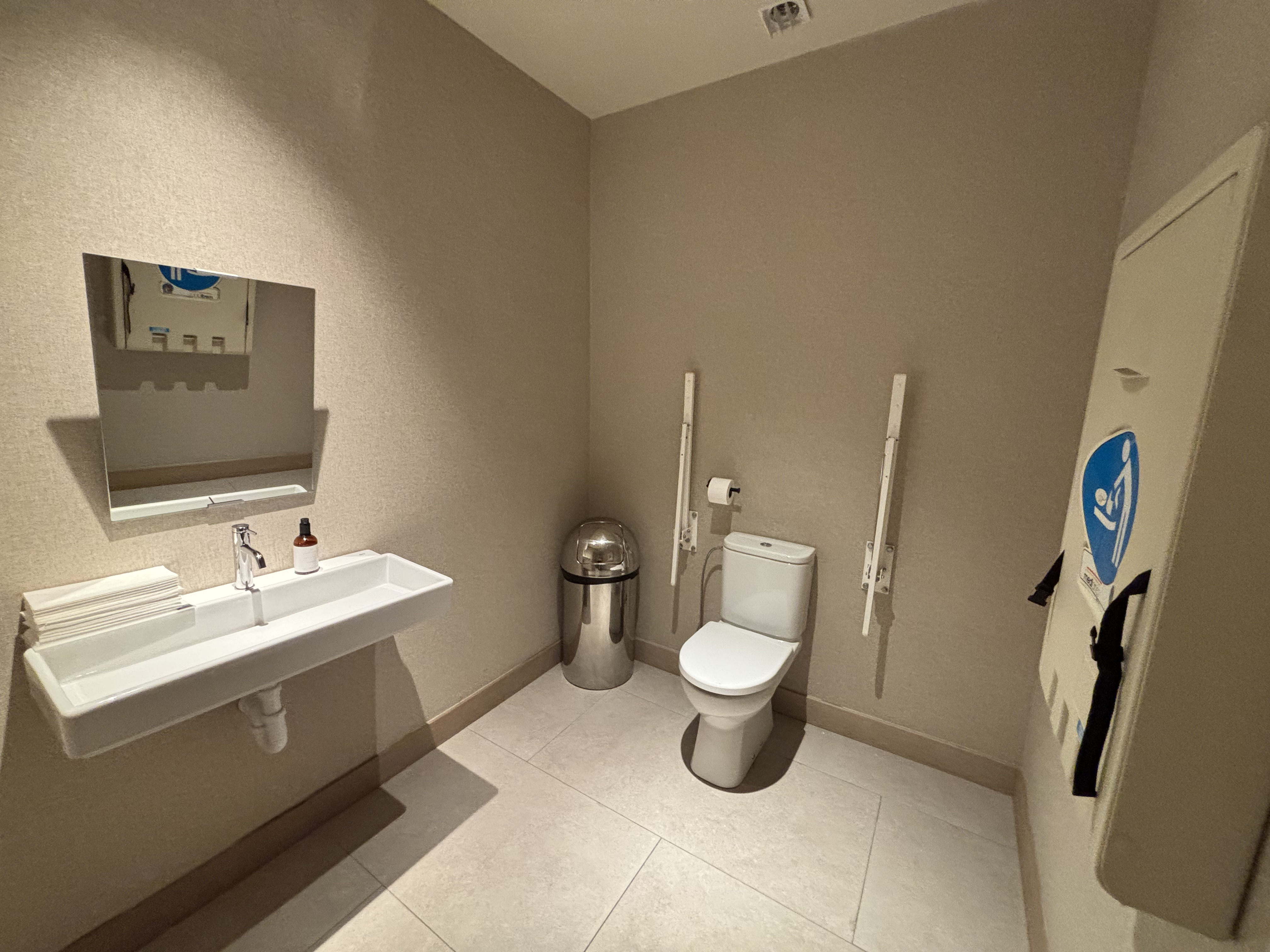 L'image montre une salle de bain accessible. À gauche, on voit un lavabo blanc avec une pile de serviettes en papier et un petit flacon dessus. Au ...