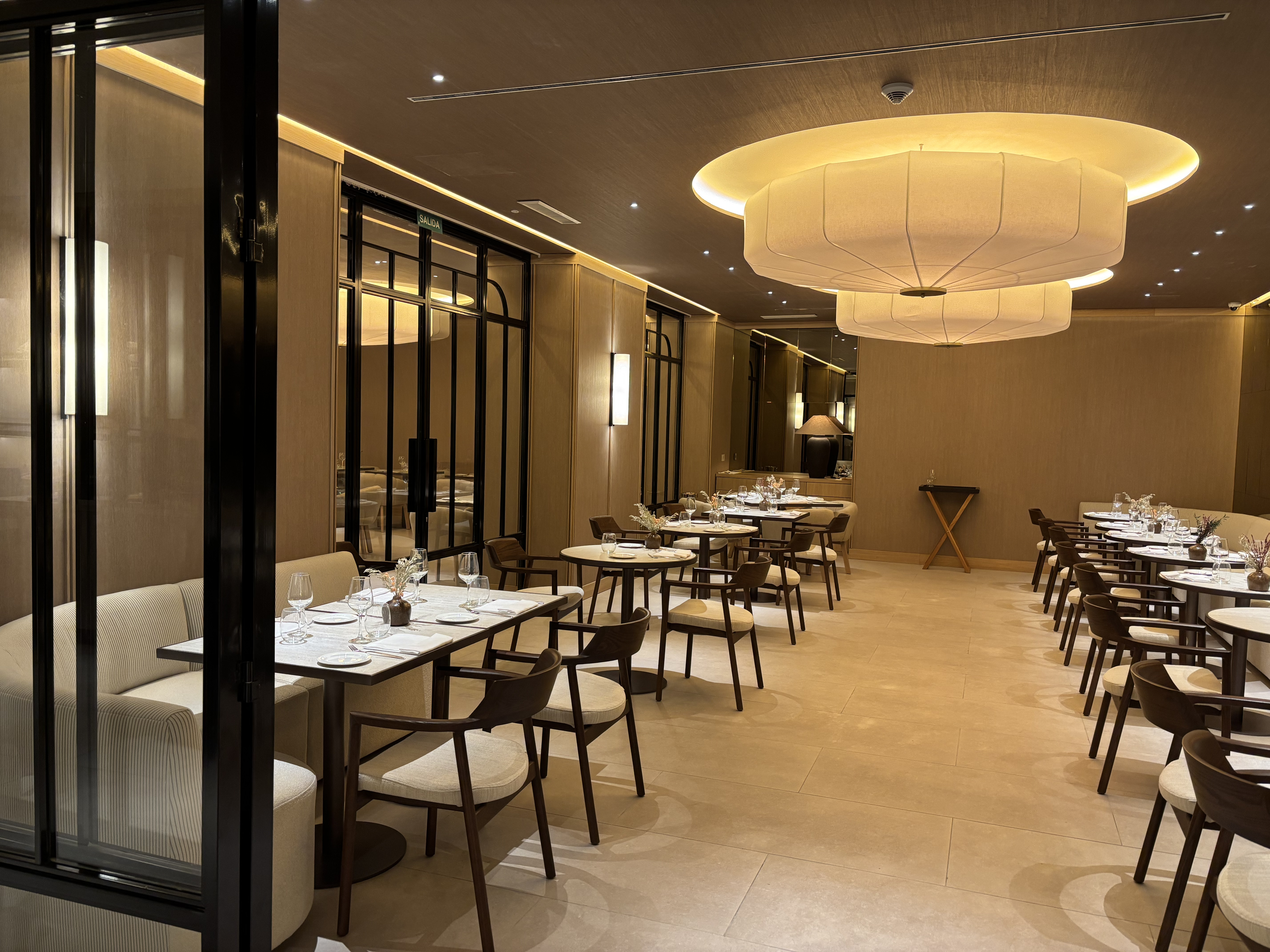 The image shows an elegant restaurant dining room. There are several round and square tables with dark wood chairs and cushioned seats. On each ...