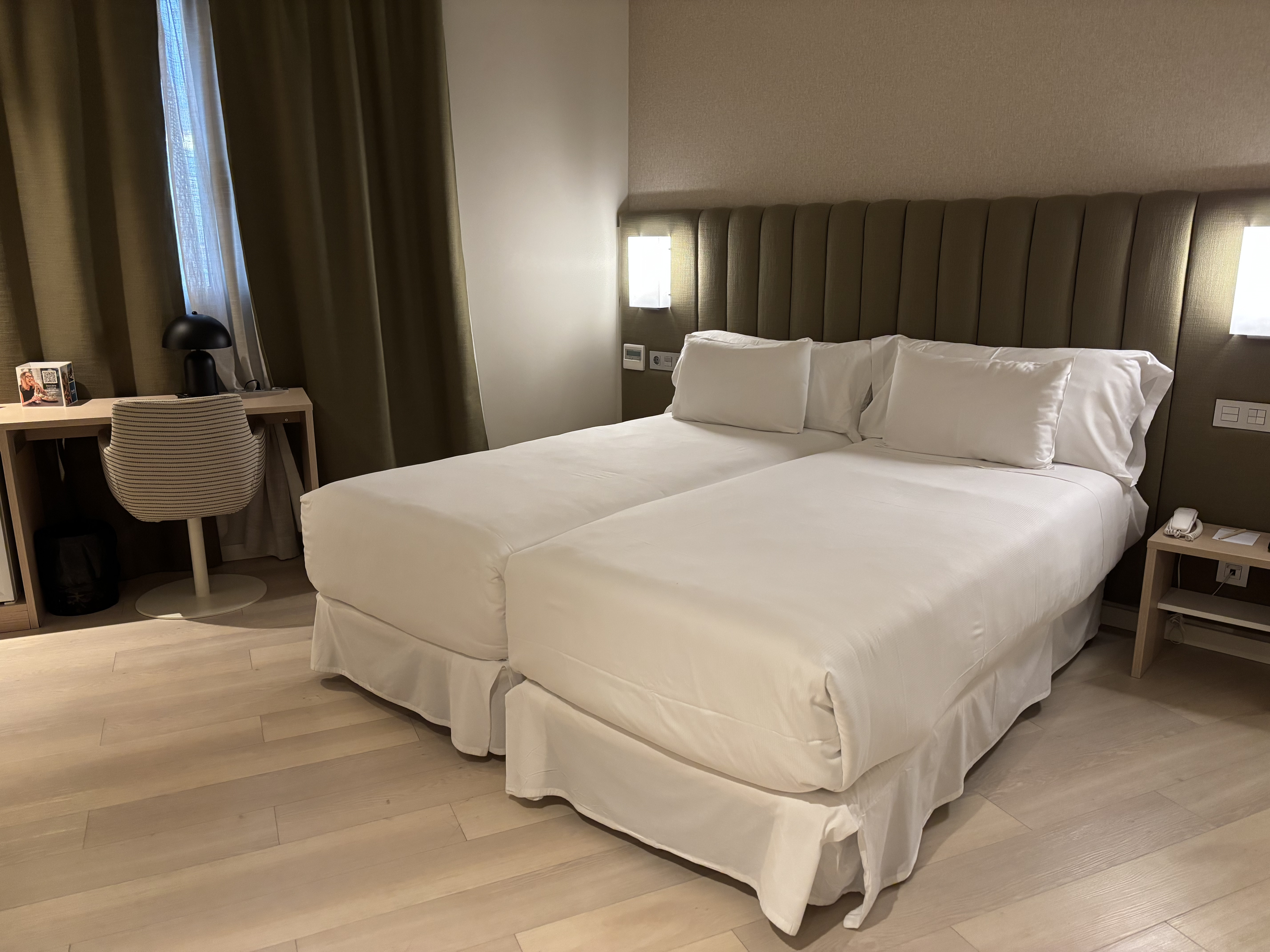 Irudiak hotel baten gela erakusten du, bi ohe bana daude. Oheak zuriak diren zapiekin estalita daude eta burko handiak dituzte. Oheen gainean, ...