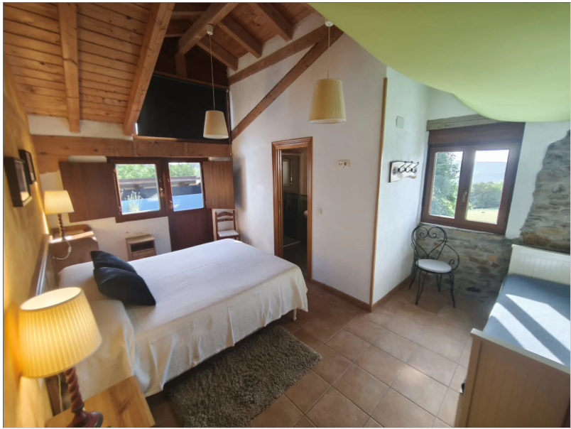 Chambre mansardée spacieuse et lumineuse, avec plafond incliné et poutres en bois apparentes. Un lit double avec du linge de lit blanc occupe le ...