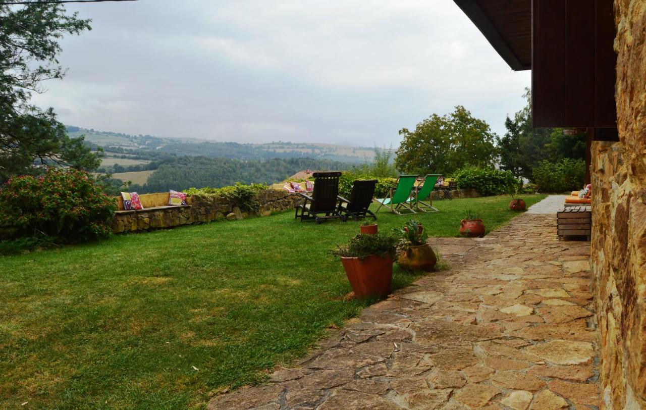 L’image montre une zone de jardin et de terrasse d’une maison rurale avec vue sur une vallée de collines vertes sous un ciel nuageux. Au premier ...