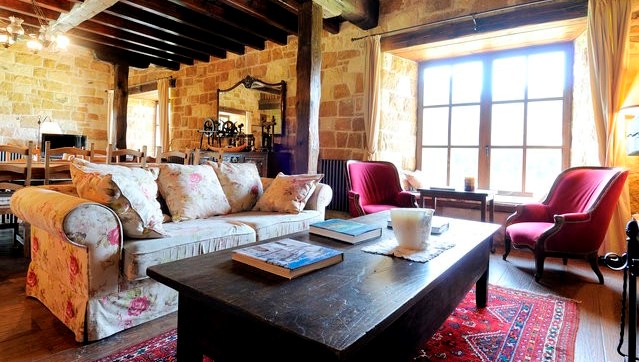 This photograph shows a rustic-style living room with stone walls and dark wooden beams on the ceiling, featuring a wooden coffee table on a red ...