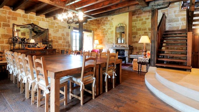 This photograph shows a rustic-style dining room with exposed stone walls and a ceiling with dark wooden beams, dominated by a very long ...